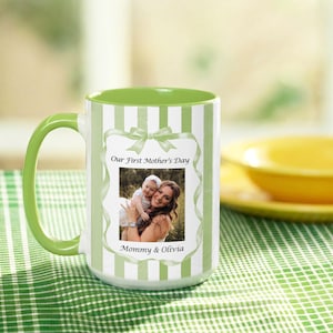 Könnte beinhalten: Weißer Keramikbecher mit grünem Griff und Innenraum. Der Becher hat vertikale grüne und weiße Streifen und ein gerahmtes Foto mit dem Text "Our First Mother's Day" und "Mommy & Olivia".