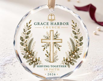 Puede incluir: Un adorno circular de cristal transparente con borde facetado, colgado de una cinta beige. Presenta una cruz dorada, ramas de hojas verdes y el texto Grace Harbor Church, Serving Together In Faith y el año 2026 impresos en oro.