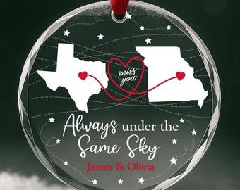 Puede incluir: Un adorno redondo de cristal transparente con una cinta roja. Muestra las siluetas blancas de Texas y Missouri conectadas por una línea roja y un corazón, con el texto "miss you", "Always under the Same Sky" y "James & Olivia" en rojo y blanco.
