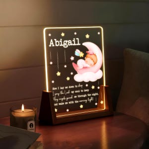 May include: A personalized night light with the name "Abigail" on a black background, featuring a sleeping baby on a crescent moon. The light has a wooden base and includes the prayer "Now I lay me down to sleep..."