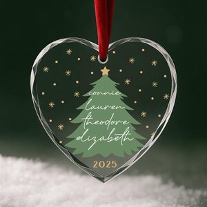 May include: Heart-shaped Christmas ornament with a red ribbon. The clear glass ornament features a green Christmas tree design with names and the year 2025. Gold stars and dots are scattered around the tree.