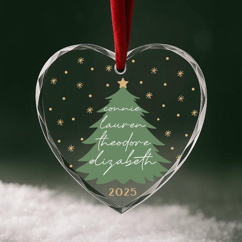 May include: Heart-shaped Christmas ornament with a red ribbon. The clear glass ornament features a green Christmas tree design with names and the year 2025. Gold stars and dots are scattered around the tree.