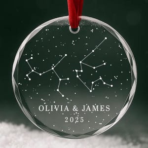 May include: A clear, round ornament with a faceted edge, featuring a constellation design and the names "OLIVIA & JAMES" with the year "2025". A red ribbon is attached for hanging. The background is a dark green.