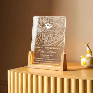May include: A clear acrylic map of the University of Michigan, engraved with a map and text. The text reads "University of Michigan, Riley Morgan, Bachelor of Science, Class of 2026." The acrylic piece is set in a wooden base with a warm light.