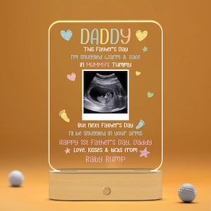 May include: A light-up acrylic sign with a wooden base. The sign features a sonogram image and the text "DADDY This Father's Day I'm snuggled warm & safe in MUMMY'S TUMMY But next Father's Day I'll be snuggled in your arms Happy 1st Father's Day, Daddy ★ Love. Kisses & kicks from Baby Bump"