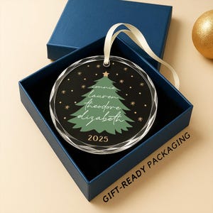 May include: A round Christmas ornament with a green tree design and names, inside a blue gift box. The ornament has a clear, faceted edge and a cream-colored ribbon for hanging. The year 2025 is printed on the ornament.