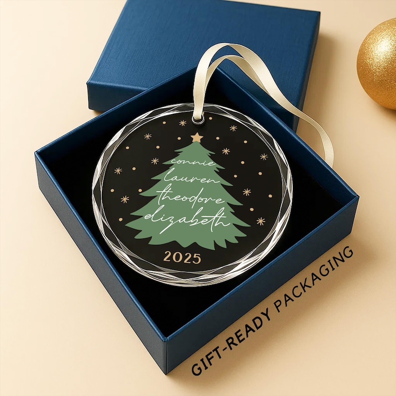 May include: A round Christmas ornament with a green tree design and names, inside a blue gift box. The ornament has a clear, faceted edge and a cream-colored ribbon for hanging. The year 2025 is printed on the ornament.