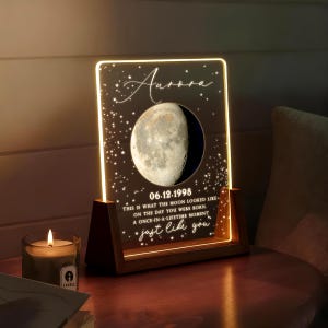 Puede incluir: Luz de noche personalizada con base de madera. El panel acrílico muestra una imagen de fase lunar, el nombre "Aurora", la fecha "06.12.1998" y el texto "This is what the moon looked like...just like you". Una vela encendida está en primer plano.
