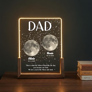 May include: A personalized LED night light with a wooden base. The clear acrylic panel displays the word "DAD" above two moon illustrations and names with dates. The text reads "This is what the Moon looked like the day you became our Dad."