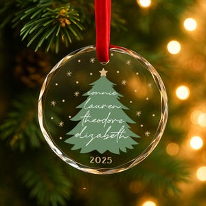 May include: A clear, round Christmas ornament with a red ribbon. The ornament features a green Christmas tree design with names and the year 2025. Snowflakes and a star are also included in the design.