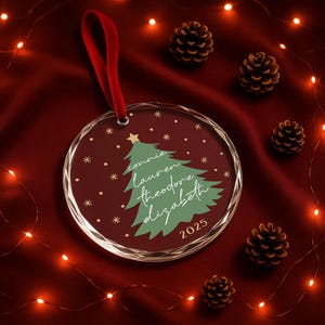 May include: A clear, round Christmas ornament with a red ribbon. The ornament features a green Christmas tree design with names and the year 2025 printed on a burgundy background. Small gold stars and dots are scattered around the tree.