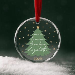 May include: A clear, round Christmas ornament with a red ribbon. The ornament features a green Christmas tree design with names and the year 2025 in white script. Gold stars and a gold star at the top of the tree complete the design.