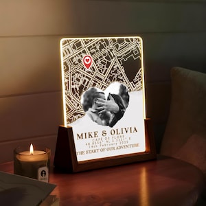 May include: A personalized illuminated map art piece with a heart-shaped photo and the names "MIKE & OLIVIA." The map shows a location with a red heart marker. Text includes "CAFE DE FLORE," coordinates, and the date "14th February 2025." The base is wooden.