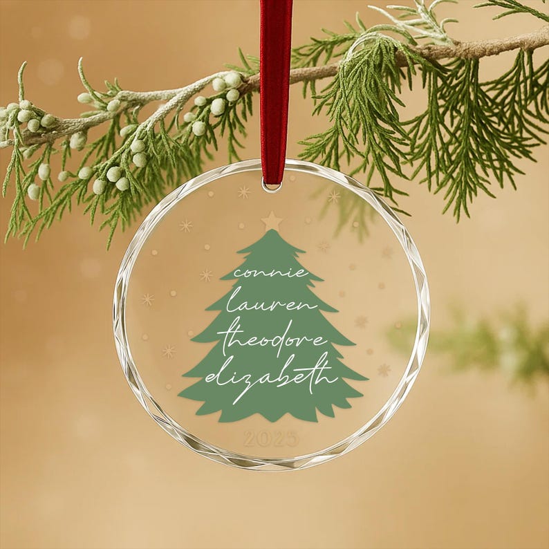 May include: A clear, round Christmas ornament with a green Christmas tree design and the names "connie, lauren, theodore, elizabeth" in white script. The ornament is suspended by a red ribbon and has the year "2023" at the bottom.