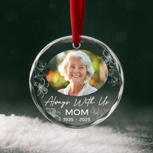 Może przedstawiać: Przezroczysta, okrągła ozdoba z fotografią uśmiechniętej kobiety, kwiatowymi akcentami i napisem "Always With Us MOM 1935-2025". Ozdoba zawieszona jest na czerwonej wstążce. Tło jest rozmyte zielone.