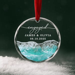 May include: Clear, round ornament with a red ribbon. The ornament has the word "engaged" in script, with the names "JAMES & OLIVIA" and the date "08.10.2025" below. The bottom of the ornament features a turquoise ocean scene.