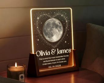 Op de afbeelding: Een rechthoekig acryl nachtlampje met houten voet, voorzien van een gloeiende maanafbeelding binnen een hart van sterren. De tekst luidt: Olivia & James, This is what the moon looked like the day we found forever in each other. Under every moon, I'll choose you. 02.14.2026.