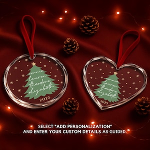 May include: Two clear Christmas ornaments with red ribbon hangers. One is round, the other heart-shaped. Both feature a green Christmas tree design, names, and the year 2025. The background is a deep red with pinecones and string lights.