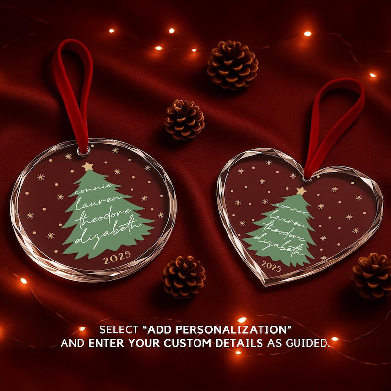 May include: Two clear Christmas ornaments with red ribbon hangers. One is round, the other heart-shaped. Both feature a green Christmas tree design, names, and the year 2025. The background is a deep red with pinecones and string lights.