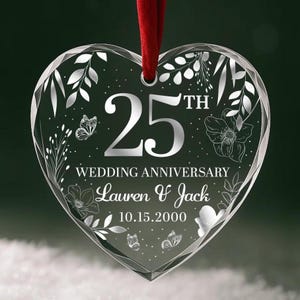 May include: A heart-shaped, clear glass ornament with a red ribbon. The ornament features the text "25th Wedding Anniversary" with the names "Lauren & Jack" and the date "10.15.2000". Decorative floral and butterfly designs are etched around the edges.