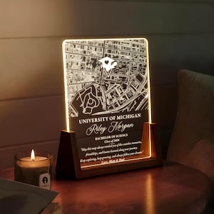 May include: A lighted map of the University of Michigan, engraved on a clear acrylic panel. The map is set in a wooden frame. Text includes the university name, a name, degree, class year, and a quote from family.