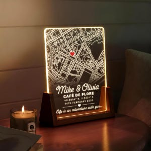 May include: A personalized illuminated map of a location, with a red heart marking a spot. The map is etched on a clear acrylic panel, set in a wooden base. Text includes names, a cafe name, coordinates, and a date.