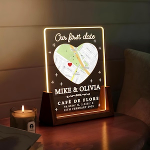 May include: A personalized LED night light with a wooden base. The acrylic panel displays a map within a heart shape, the text "Our first date", names, location, and date. A lit candle sits nearby.