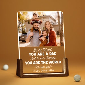 May include: A photo of a family of four, a father, mother, and two young boys, in front of a house. The photo is on a clear acrylic plaque with a wooden base. The text on the plaque reads: "To the World YOU ARE A DAD But to our Family YOU ARE THE WORLD -We love you! - Cindy, Olivia, Mike"