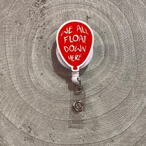 Könnte beinhalten: Roter Ballon mit dem Text "We All Float Down Here" auf weißem Hintergrund. Der Ballon ist an einem durchsichtigen, einziehbaren Ausweishalter aus Kunststoff befestigt.
