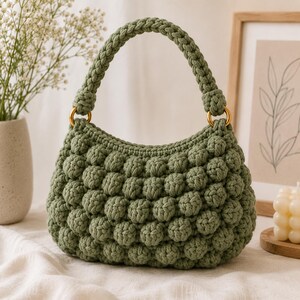 Peut inclure: Un sac à main en crochet vert sauge présentant un motif texturé en points noisette sur tout le corps. Le sac possède une anse tressée unique fixée au compartiment principal par deux anneaux métalliques dorés, formant une silhouette arrondie.