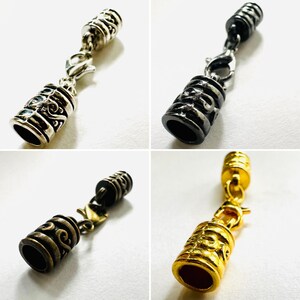 May include: Four metal pendant charms in silver, black, bronze, and gold. Each charm features a cylindrical barrel design with ornate detailing and a clasp for attachment. The charms are displayed against a white background, showcasing their different finishes.