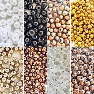 May include: A selection of small, round beads in diverse colours, such as white, black, gold, and bronze. The beads have a shiny, metallic finish and are closely packed together. Ideal for jewellery making and crafts, these beads offer a range of creative possibilities.