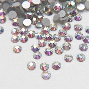 May include: A collection of iridescent rhinestones scattered on a white surface. The rhinestones are round with a flat, silver backing and a faceted, multi-colored top that reflects pink, purple, and blue hues. Ideal for crafting and embellishments.