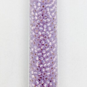 May include: A clear tube filled with small, light purple seed beads. The beads have a slightly iridescent quality, reflecting light. These beads are cylindrical with a small hole running through the center, perfect for stringing.