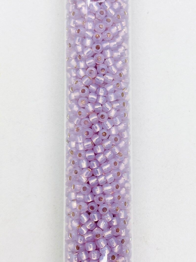 May include: A clear tube filled with small, light purple seed beads. The beads have a slightly iridescent quality, reflecting light. These beads are cylindrical with a small hole running through the center, perfect for stringing.