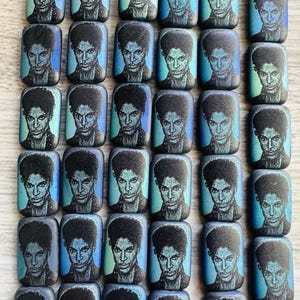 May include: Rectangular pendants featuring a portrait of a musician with dark hair and a purple jacket. The pendants have a blue and green background. The pendants are strung together with a thin, clear string.