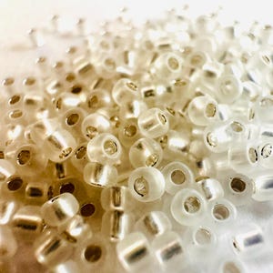 May include: A close-up shot of a pile of clear, cylindrical beads with silver-colored centers. The beads are scattered, creating a textured surface. The lighting highlights the translucent quality of the beads, giving them a subtle shimmer. Ideal for jewelry making.