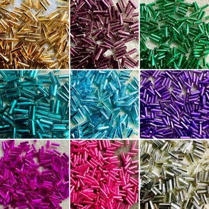 May include: A collection of nine distinct piles of small, cylindrical glass bugle beads in various metallic and vibrant colors, including gold, dark purple, emerald green, teal, light blue, violet, magenta, hot pink, and silver.