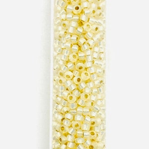 May include: A clear tube filled with small, pale yellow seed beads. The beads have a slightly iridescent quality, reflecting light. These beads are ideal for jewelry making and crafting projects.