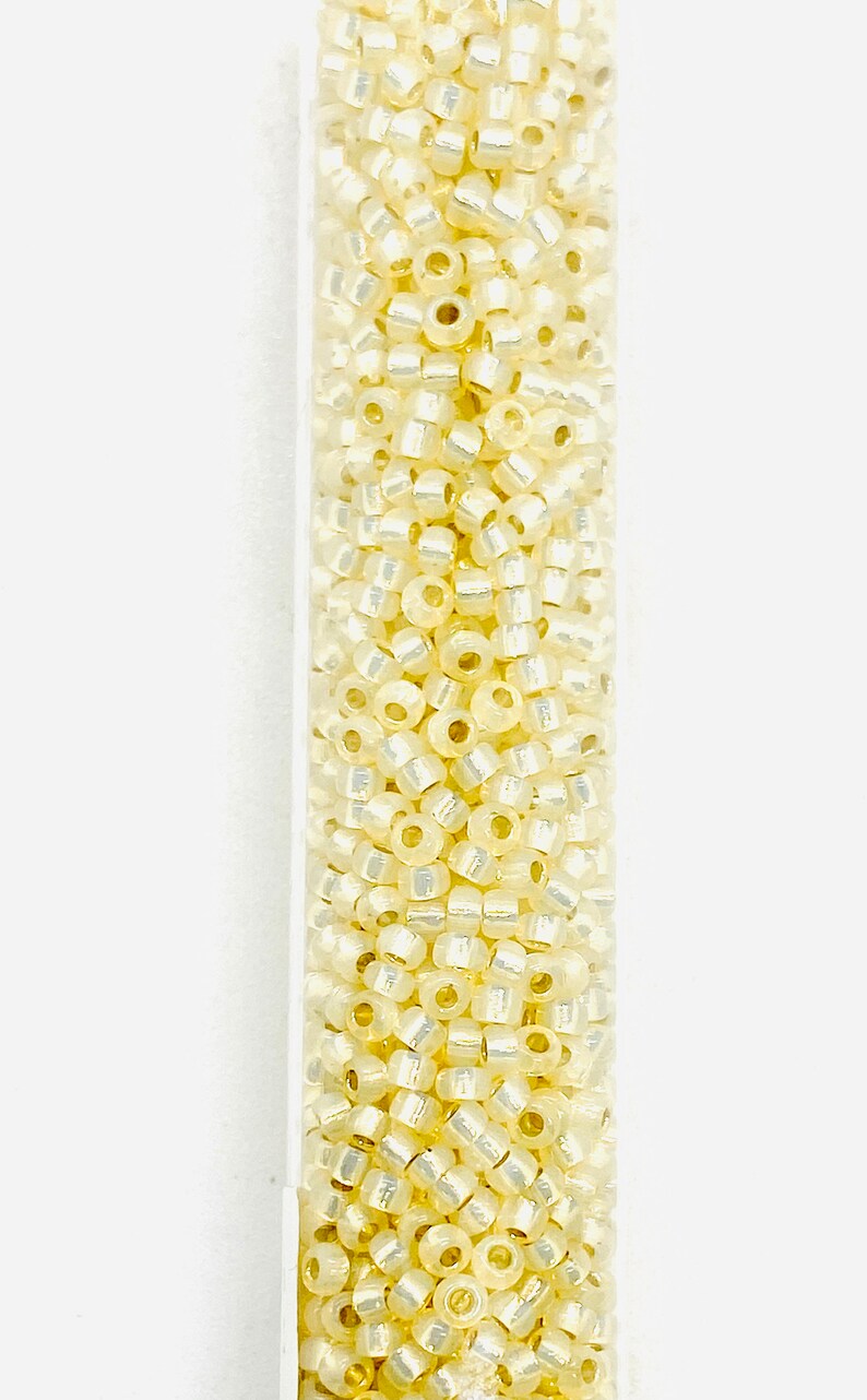 May include: A clear tube filled with small, pale yellow seed beads. The beads have a slightly iridescent quality, reflecting light. These beads are ideal for jewelry making and crafting projects.
