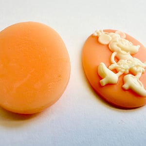 May include: Two oval-shaped cameo pendants. One is plain peach-colored, the other features a cream-colored Minnie Mouse design on a peach background. The cameo is a classic jewelry element, often used in necklaces, bracelets, and earrings.