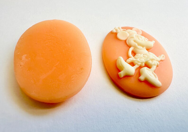 May include: Two oval-shaped cameo pendants. One is plain peach-colored, the other features a cream-colored Minnie Mouse design on a peach background. The cameo is a classic jewelry element, often used in necklaces, bracelets, and earrings.