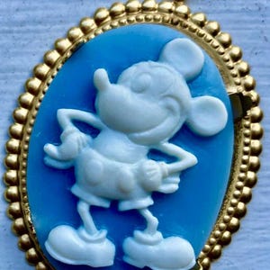 May include: Oval cameo pendant featuring a white Mickey Mouse figure on a light blue background, set within a gold-colored frame. The frame has a beaded edge and a loop at the top for hanging. A classic Disney-themed accessory.