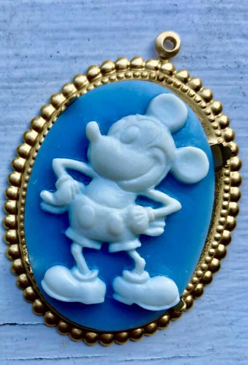 May include: Oval cameo pendant featuring a white Mickey Mouse figure on a light blue background, set within a gold-colored frame. The frame has a beaded edge and a loop at the top for hanging. A classic Disney-themed accessory.