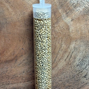 May include: A clear plastic tube filled with small, round, gold-colored beads. The tube has a white lid with a loop handle.