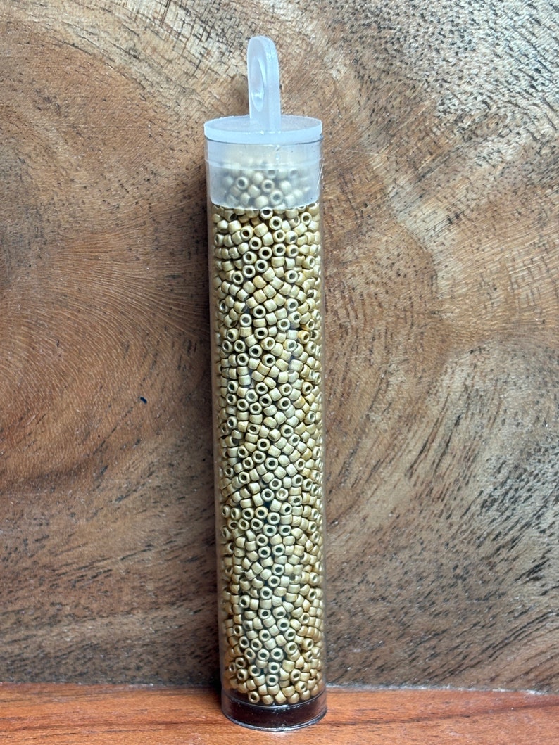 May include: A clear plastic tube filled with small, round, gold-colored beads. The tube has a white lid with a loop handle.