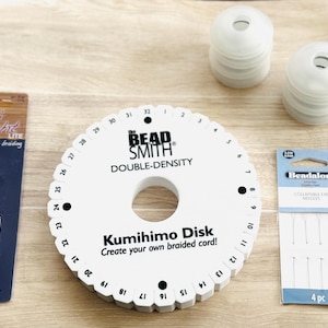 May include: A collection of crafting supplies for Kumihimo braiding. Includes a white double-density Kumihimo disk, a silver weight, a package of needles, and two stacks of white spools. The disk has the text "The Bead Smith Double-Density Kumihimo Disk".
