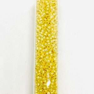 May include: A clear tube filled with small, yellow seed beads. The beads have a slightly iridescent quality, reflecting light. The tube is vertical against a white background. These beads are suitable for jewelry making and craft projects.