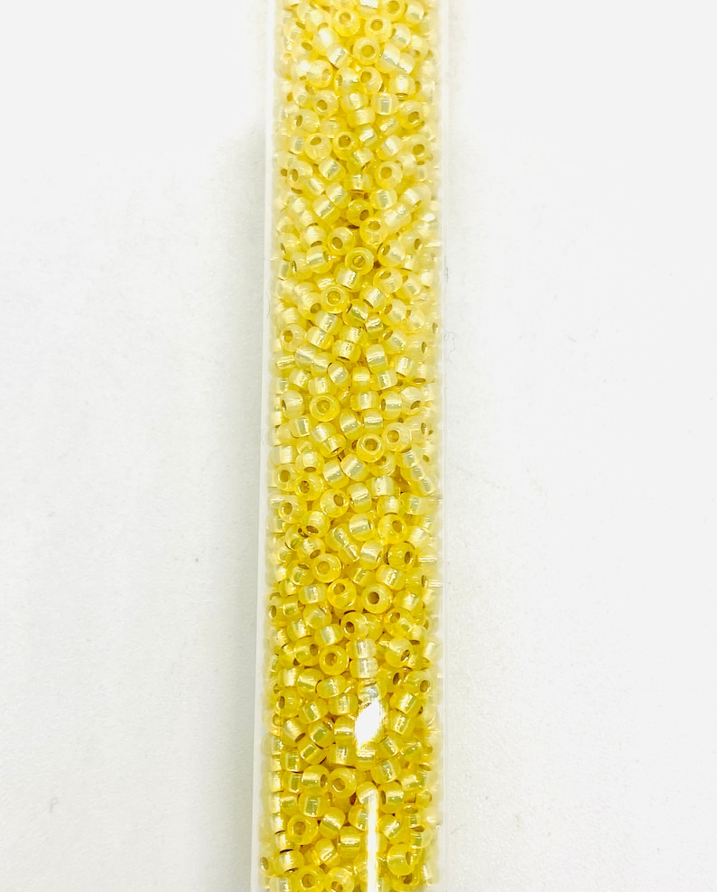May include: A clear tube filled with small, yellow seed beads. The beads have a slightly iridescent quality, reflecting light. The tube is vertical against a white background. These beads are suitable for jewelry making and craft projects.