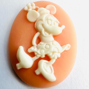 May include: A cameo featuring a cream-colored Minnie Mouse figure on a peach-colored oval background. Minnie Mouse is depicted in a classic pose, wearing a polka-dot skirt and a bow. The cameo is likely made of resin or a similar material.